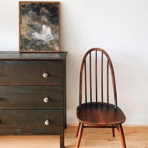 May include: A dark wood dresser with three drawers and black ceramic knobs. A framed print of a girl in a white dress sitting in a field of flowers hangs above the dresser. A wooden chair with a curved back and spindle legs sits to the right of the dresser.
