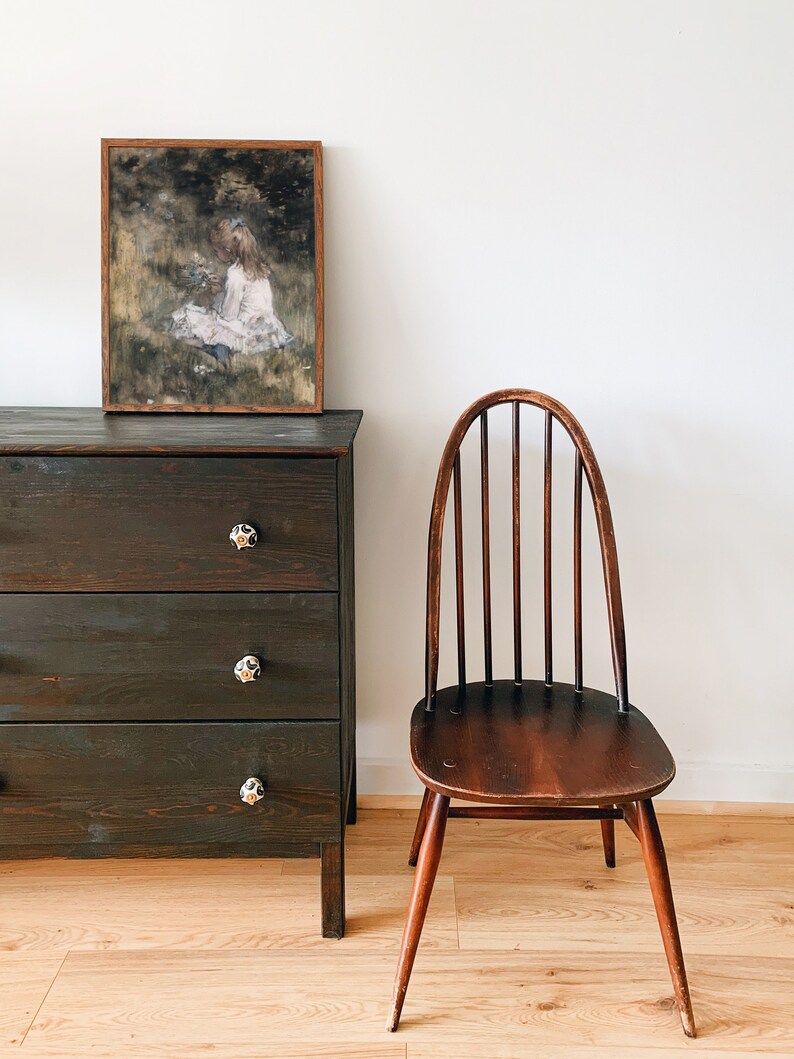 May include: A dark wood dresser with three drawers and black ceramic knobs. A framed print of a girl in a white dress sitting in a field of flowers hangs above the dresser. A wooden chair with a curved back and spindle legs sits to the right of the dresser.