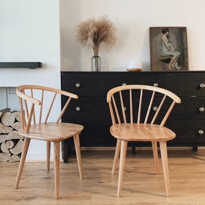 May include: Two light wood chairs with a spindle back design, sitting in front of a black dresser with a painting above it.