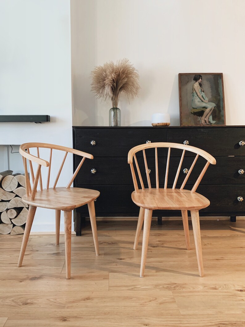 May include: Two light wood chairs with a spindle back design, sitting in front of a black dresser with a painting above it.