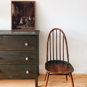 May include: A dark wood dresser with three drawers and black knobs. A framed picture of a group of children in a room hangs above the dresser. A wooden chair with a curved back and a dark wood finish sits to the right of the dresser.