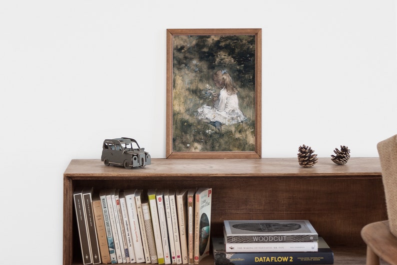 May include: A framed painting of a young girl sitting in a field of flowers. The painting is in a wooden frame and is hanging on a wall above a shelf with books and a toy car.