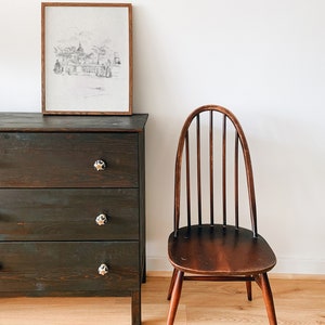 May include: A dark wood dresser with three drawers and white ceramic knobs. A framed black and white drawing of a city scene hangs above the dresser. A wooden chair with a curved back and spindle back sits on a light wood floor.