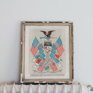 May include: A framed vintage print of an American eagle with a collection of flags surrounding it. The flags are red, white, and blue and include the American flag, the Confederate flag, and other historical flags. The print is in a silver frame and is hanging on a white wall.