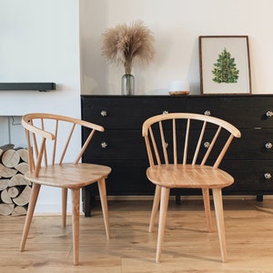 May include: Two light wood chairs with a spindle back design sit in front of a black dresser with a framed picture of a green Christmas tree above it.