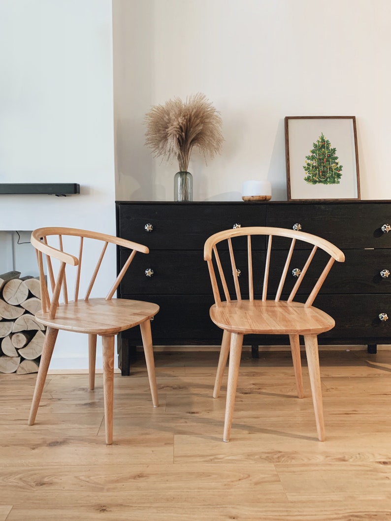 May include: Two light wood chairs with a spindle back design sit in front of a black dresser with a framed picture of a green Christmas tree above it.