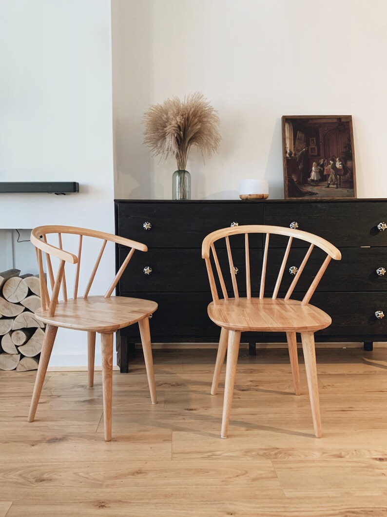 May include: Two light wood chairs with a spindle back and a curved seat. The chairs are in front of a black dresser with a picture hanging above it.