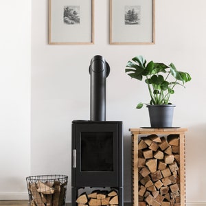 May include: A black wood-burning stove with a black pipe and a glass door. The stove is sitting on a white tile floor. There is a basket of firewood next to the stove and a wooden stand with a stack of firewood next to it. There is a potted plant with green leaves on the stand. Two framed black and white prints are hanging on the white wall behind the stove.
