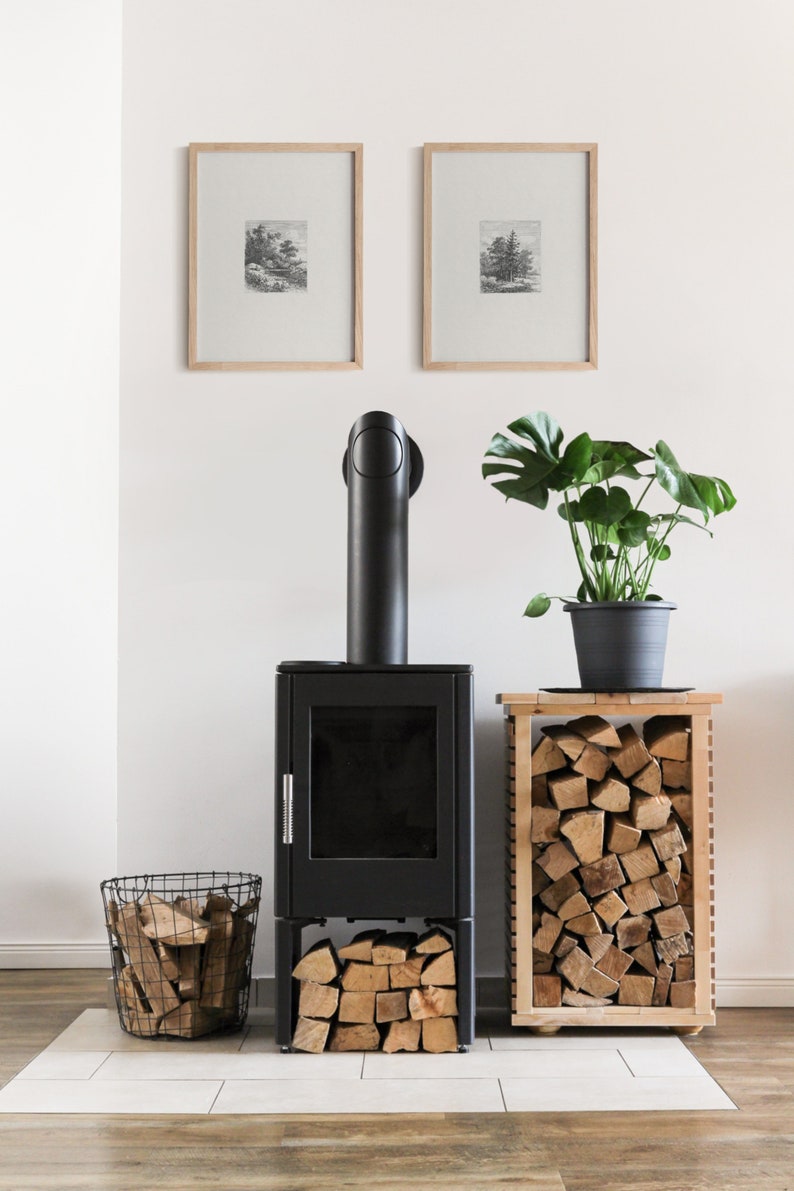 May include: A black wood-burning stove with a black pipe and a glass door. The stove is sitting on a white tile floor. There is a basket of firewood next to the stove and a wooden stand with a stack of firewood next to it. There is a potted plant with green leaves on the stand. Two framed black and white prints are hanging on the white wall behind the stove.