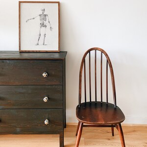 May include: A dark wood dresser with three drawers and black ceramic knobs. A wooden chair with a curved back and a dark wood finish sits to the right of the dresser. A framed drawing of a human skeleton hangs on the wall above the dresser.
