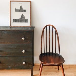 Peut inclure: Une commode en bois fonc&eacute; avec trois tiroirs et des boutons noirs. Une estampe en noir et blanc encadr&eacute;e d'un dessin architectural d'un b&acirc;timent est accroch&eacute;e au-dessus de la commode. Une chaise en bois avec un dossier &agrave; lattes et un si&egrave;ge brun est plac&eacute;e &agrave; droite de la commode.