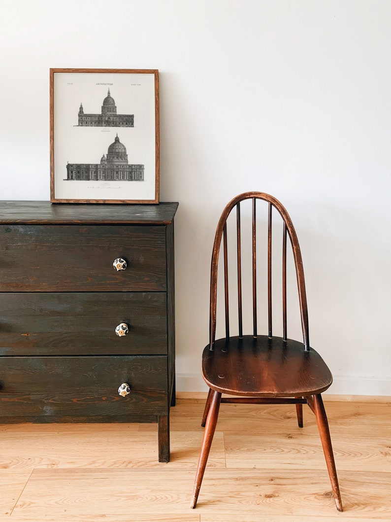 Peut inclure: Une commode en bois fonc&eacute; avec trois tiroirs et des boutons noirs. Une estampe en noir et blanc encadr&eacute;e d'un dessin architectural d'un b&acirc;timent est accroch&eacute;e au-dessus de la commode. Une chaise en bois avec un dossier &agrave; lattes et un si&egrave;ge brun est plac&eacute;e &agrave; droite de la commode.
