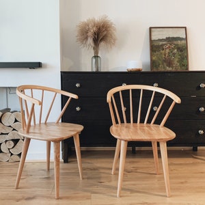 May include: Two light wood chairs with a spindle back design and a rounded seat. The chairs are in front of a black dresser with a painting above it.