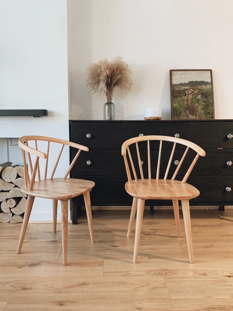 May include: Two light wood chairs with a spindle back design and a rounded seat. The chairs are in front of a black dresser with a painting above it.