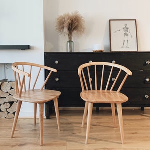 May include: Two light wood chairs with a spindle back design and a rounded seat. The chairs are in front of a black dresser with drawers.