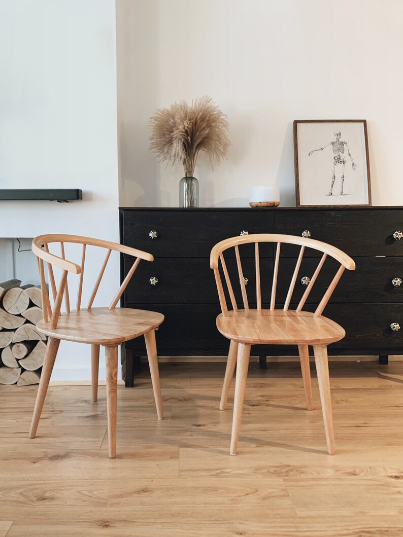 May include: Two light wood chairs with a spindle back design and a rounded seat. The chairs are in front of a black dresser with drawers.