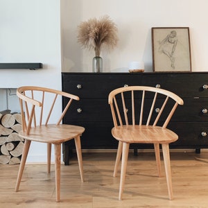 May include: Two light wood chairs with a spindle back and a curved seat. The chairs are in front of a black dresser with a vase of dried pampas grass on top.
