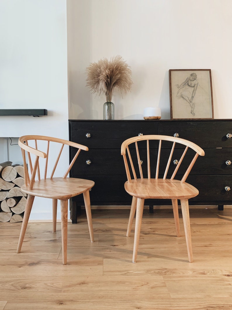 May include: Two light wood chairs with a spindle back and a curved seat. The chairs are in front of a black dresser with a vase of dried pampas grass on top.