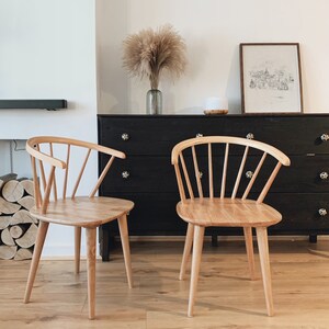 May include: Two light wood chairs with a spindle back design sit in front of a black dresser with a vase of dried pampas grass on top.