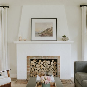 May include: A white fireplace with a brick surround and a wood-burning fire. The fireplace is decorated with a framed landscape painting and a potted plant.