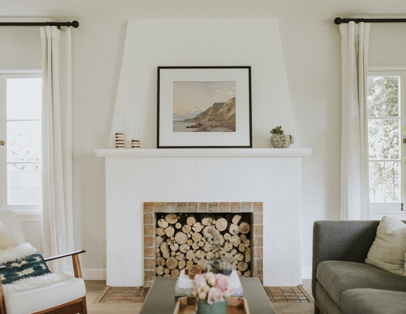 May include: A white fireplace with a brick surround and a wood-burning fire. The fireplace is decorated with a framed landscape painting and a potted plant.