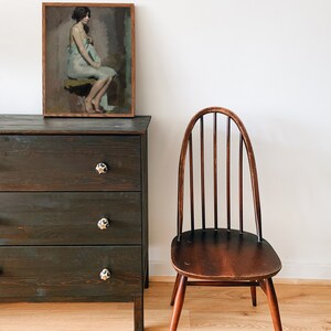 May include: A dark wood dresser with three drawers and black ceramic knobs. The dresser has a framed painting of a woman sitting on a stool, wearing a light blue dress. A dark wood chair with a curved back and spindle back is in the foreground.