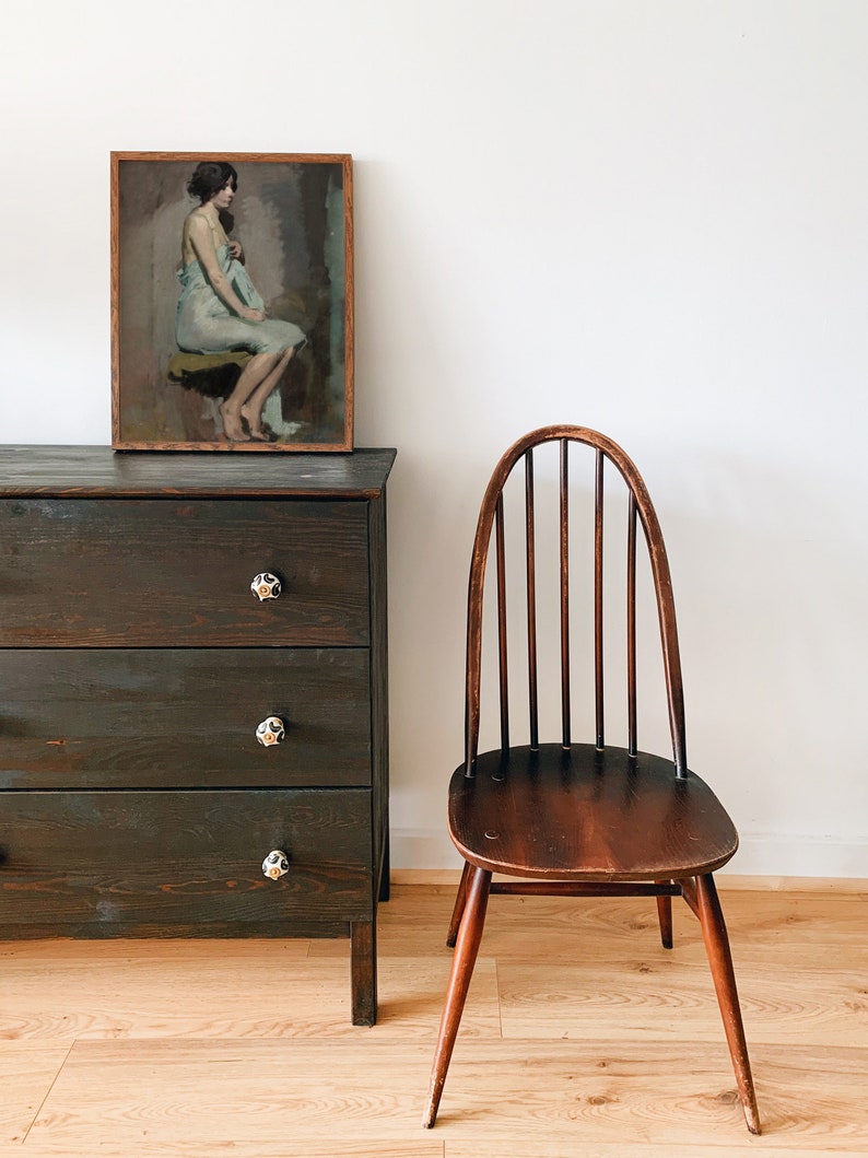 May include: A dark wood dresser with three drawers and black ceramic knobs. The dresser has a framed painting of a woman sitting on a stool, wearing a light blue dress. A dark wood chair with a curved back and spindle back is in the foreground.