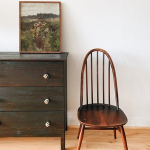 May include: A dark wood dresser with three drawers and black knobs. A framed landscape painting hangs above the dresser. A wooden chair with a curved back and spindles sits to the right of the dresser. The chair and dresser are in front of a white wall.