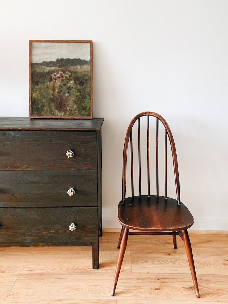May include: A dark wood dresser with three drawers and black knobs. A framed landscape painting hangs above the dresser. A wooden chair with a curved back and spindles sits to the right of the dresser. The chair and dresser are in front of a white wall.