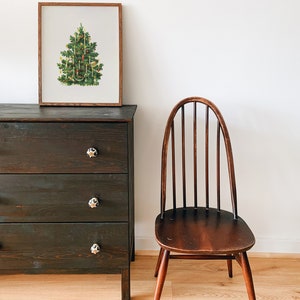 May include: A framed illustration of a Christmas tree decorated with ornaments and candles, a dark wooden dresser with three drawers, and a wooden chair with a spindle back.