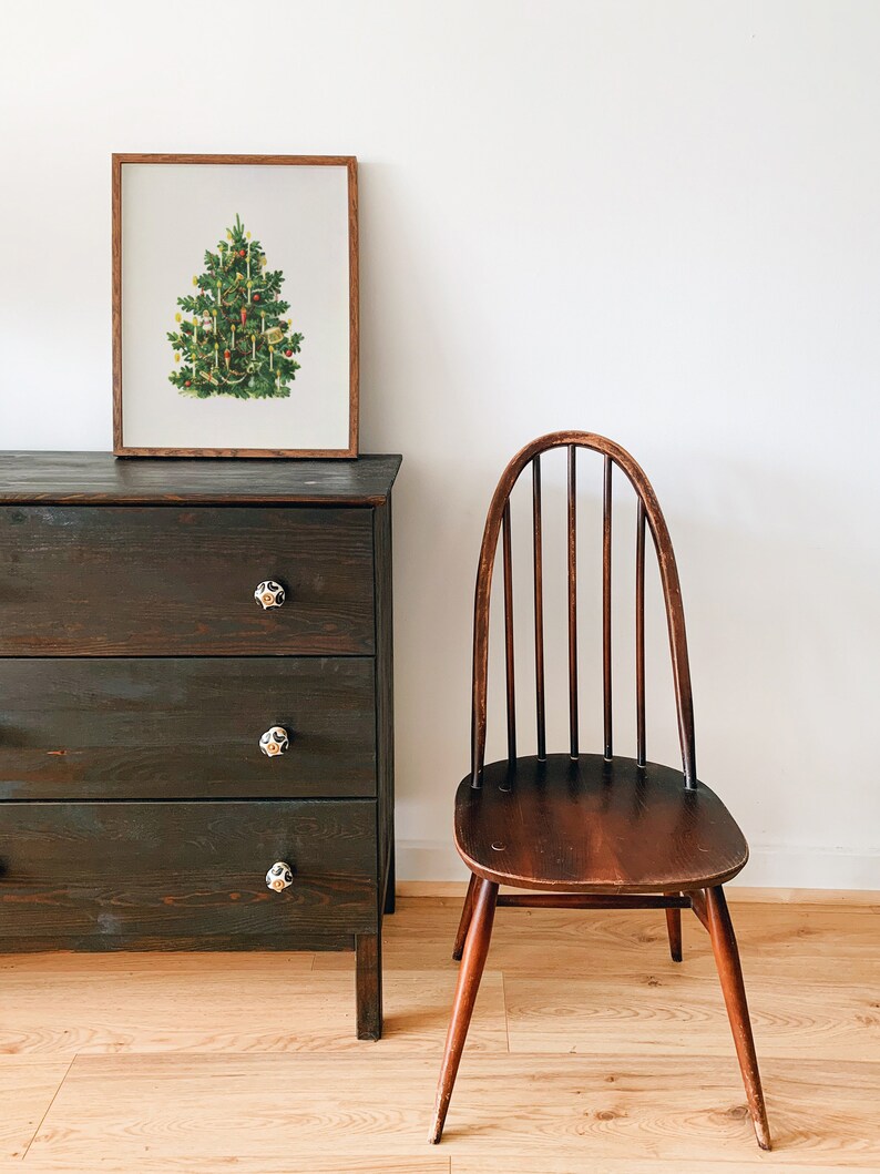 May include: A framed illustration of a Christmas tree decorated with ornaments and candles, a dark wooden dresser with three drawers, and a wooden chair with a spindle back.