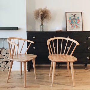 May include: Two light wood chairs with a spindle back design sit in front of a black dresser with a framed print of an eagle and flags above it.