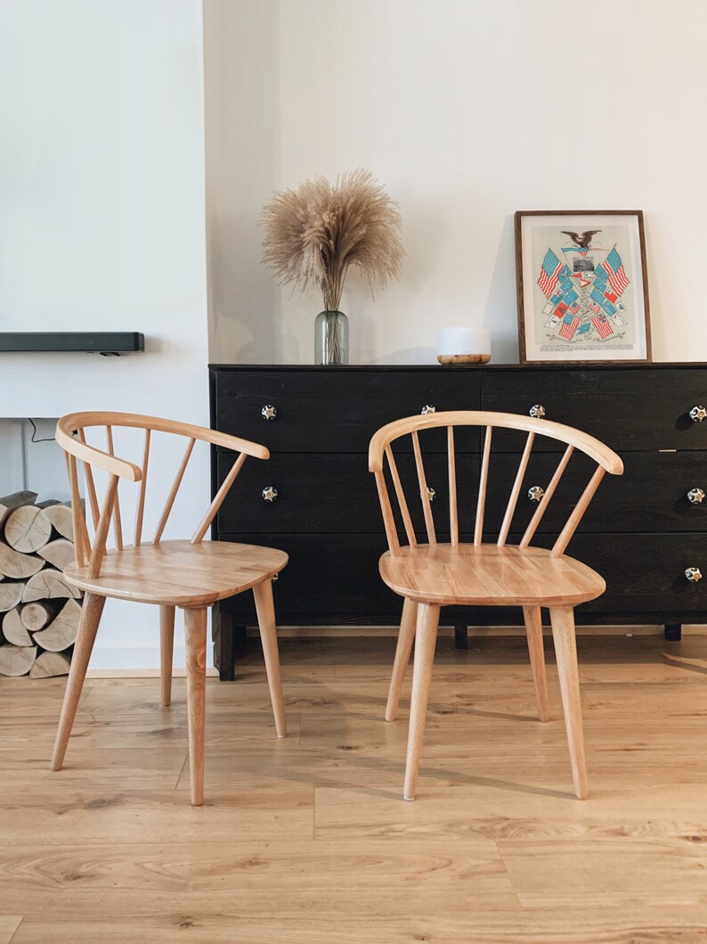 May include: Two light wood chairs with a spindle back design sit in front of a black dresser with a framed print of an eagle and flags above it.