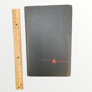 May include: A black book with a red star and line design. The book is approximately 9 inches tall, as measured by the wooden ruler on the left. The book's cover is slightly worn.