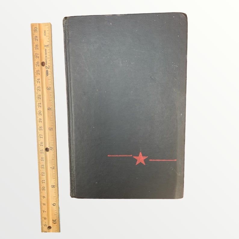 May include: A black book with a red star and line design. The book is approximately 9 inches tall, as measured by the wooden ruler on the left. The book's cover is slightly worn.