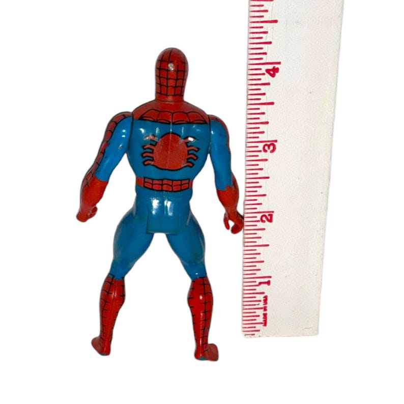 May include: A vintage Spider-Man action figure, featuring a red and blue color scheme. The figure is approximately 4 inches tall, with a red spider emblem on the back. The figure is standing next to a ruler.