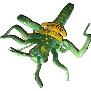 Peut inclure: Un jouet insecte en plastique vert avec des yeux rouges et des taches jaunes sur le dos. Il comporte une étiquette jaune attachée au corps avec l'inscription SUPER DUPER WEIRD WIGGLIE, MADE IN HONG KONG. Le jouet a plusieurs pattes articulées.