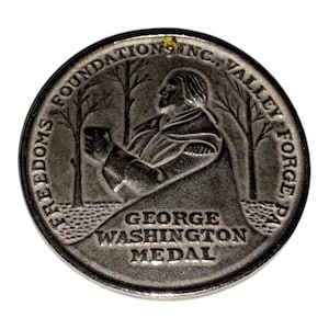 May include: A circular silver-toned metal medal featuring a relief profile of George Washington standing between two trees. The text reads FREEDOMS FOUNDATION INC., VALLEY FORGE, PA along the top edge and GEORGE WASHINGTON MEDAL at the bottom.