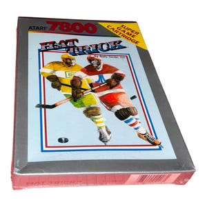 Op de afbeelding: Atari 7800 Hat Trick Super Game Cartridge speldoos. De hoes toont twee hockeyspelers in gele en rode shirts met sticks op het ijs. De tekst bevat Atari 7800, Super Game Cartridge, Hat Trick en by Bally Sente, Inc. De doos is zilverkleurig.