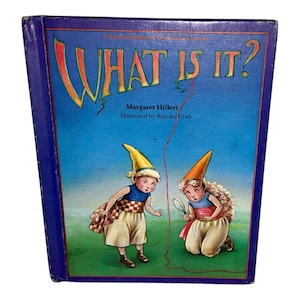 May include: A children's book titled "WHAT IS IT?" with a blue cover. The cover features illustrations of two cartoon characters in elf-like costumes, with the title in large, orange, whimsical letters. The author is Margaret Hillert, and the illustrator is Kinuko Craft.