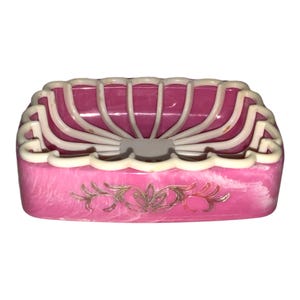 May include: A vintage pink soap dish with a white, scalloped rim and a decorative gold floral design. The dish has a fan-like design on top and a marbled effect on the base. A charming bathroom accessory.