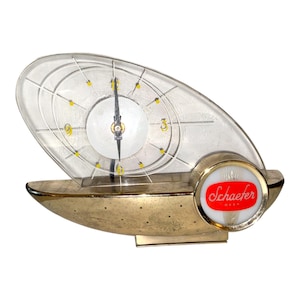 Puede incluir: Reloj dorado vintage con esfera clara de forma ovalada. El reloj presenta marcadores horarios amarillos y manecillas negras. La base tiene la palabra "Schaefer" en rojo sobre fondo blanco.