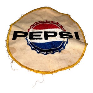 Puede incluir: Un parche vintage de Pepsi con un fondo color crema y un borde dorado. El logotipo de Pepsi está en el centro, con un diseño de tapón de botella rojo y azul con la palabra "PEPSI" en letras negras en negrita.