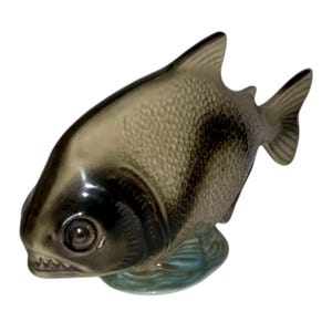 May include: A ceramic fish figurine with a detailed scale pattern in shades of gray and black. The fish has a wide mouth with visible teeth and is resting on a small, light blue base, suggesting water. The figurine is a decorative item.