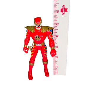 May include: A red action figure with gold and blue accents, standing next to a ruler. The figure has a gold helmet, shoulder pads, and belt buckle. The ruler shows the figure is approximately 4 inches tall.