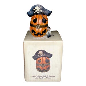 May include: A decorative orange jack-o'-lantern figurine wearing a black pirate hat with a white skull and crossbones, an eye patch, and a belt with a paw print buckle. Beside it stands a small mouse figurine. The box displays the text Pegleg's Pirate Jack-O-Lantern with Patch McNibble.