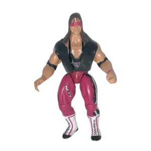 May include: A vintage wrestler action figure. The figure is wearing pink and black tights with white boots, a black vest, and pink sunglasses. The figure has long brown hair and is posed with arms outstretched.