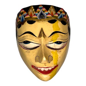 Könnte beinhalten: Eine handbemalte Holzmaske mit einem lächelnden Gesicht. Die Maske hat ein cremefarbenes Gesicht mit schwarzen Augenbrauen, Augen und Haaren. Der Mund ist rot, und die Maske ist mit einem dekorativen Kopfschmuck mit goldenen, blauen und roten Akzenten verziert.