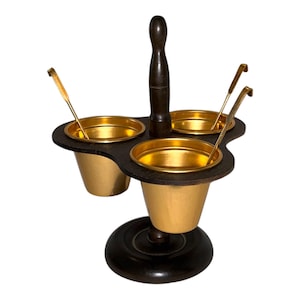 May include: A condiment server featuring three removable gold-toned metal cups held within a dark brown wooden frame. The stand includes a central vertical handle and a circular base, accompanied by three matching gold-toned metal spoons.