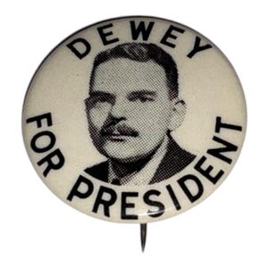 May include: A vintage campaign button with the text "DEWEY FOR PRESIDENT" encircling a black and white portrait of a man with a mustache. The button is off-white with a metal pin.